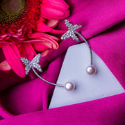 A pair of silver earcuffs featuring embellished butterfly motifs with pearl accents, displayed on a pink fabric surface.