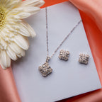 A silver cubic pendant necklace with a matching pair of earrings, displayed on a white surface with a flower in the background.