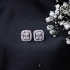 A pair of sterling silver cushion-shaped earrings with intricate detailing, displayed on a dark fabric surface alongside some small white flowers.