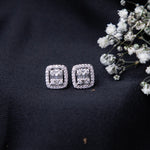 A pair of sterling silver cushion-shaped earrings with intricate detailing, displayed on a dark fabric surface alongside some small white flowers.