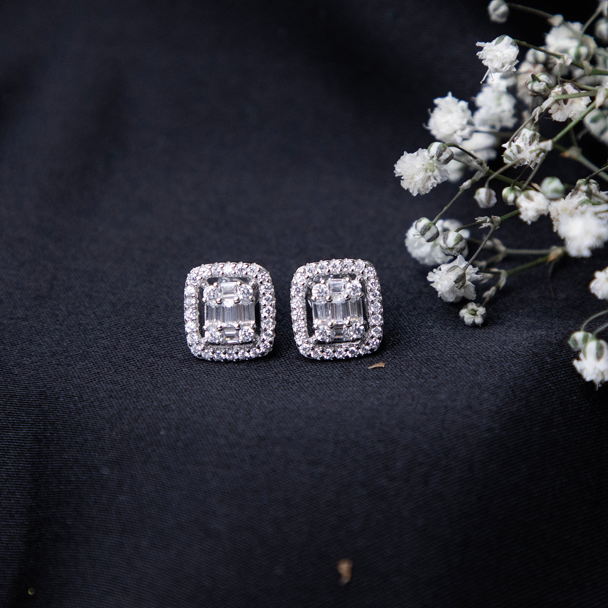 A pair of sterling silver cushion-shaped earrings with intricate detailing, displayed on a dark fabric surface alongside some small white flowers.
