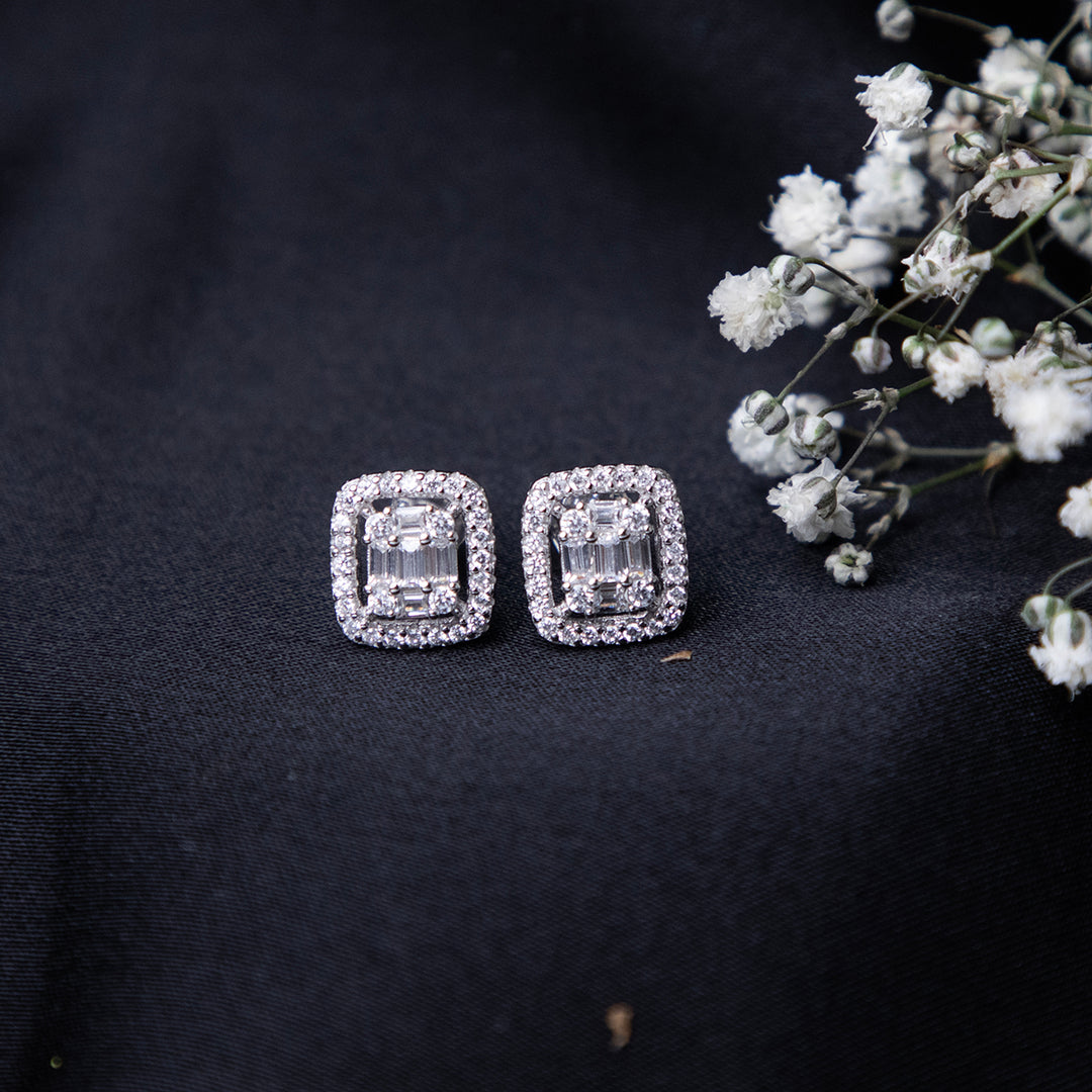 A pair of sterling silver cushion-shaped earrings with intricate detailing, displayed on a dark fabric surface alongside some small white flowers.