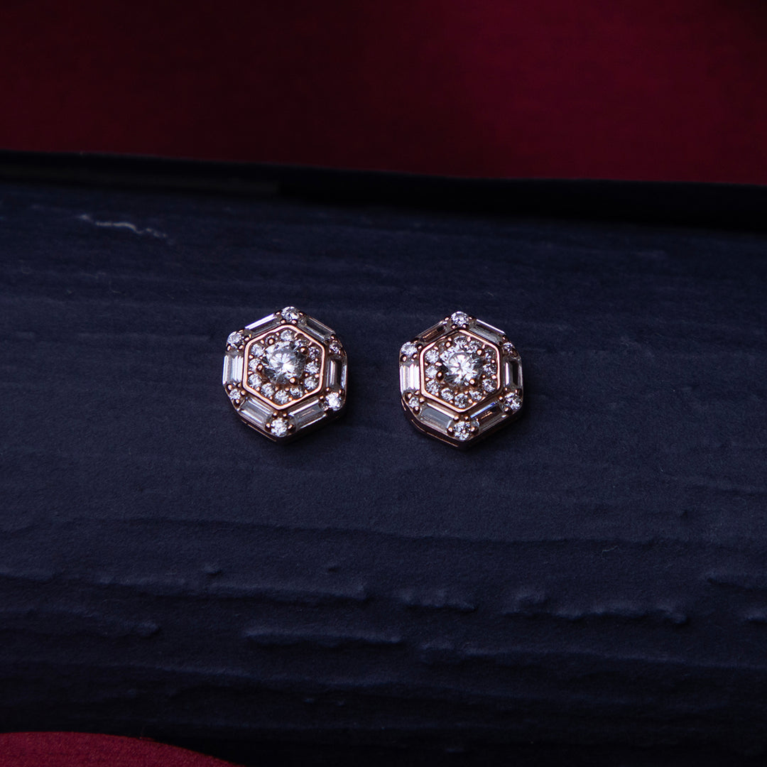 A pair of rose gold-colored sterling silver stud earrings with a halo design and a central brilliant stone, placed on a dark surface.