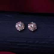 A pair of rose gold stud earrings with a halo design and a central brilliant stone, placed on a dark surface.