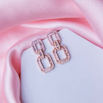 A pair of rose gold finished chain link earrings with embellishments, displayed on a pink fabric surface.