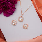 A set of rose gold colored jewelry including a pendant with a central pearl and two square earrings, displayed on a white surface with a flower and a book in the background.