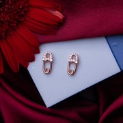 A pair of rose gold safety pin stud earrings with embellishments, displayed on a flat surface with a red flower and a blue textured background.
