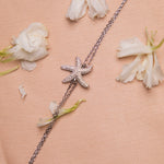 A silver bracelet featuring an embellished starfish charm, displayed on a pink background with scattered white flowers.