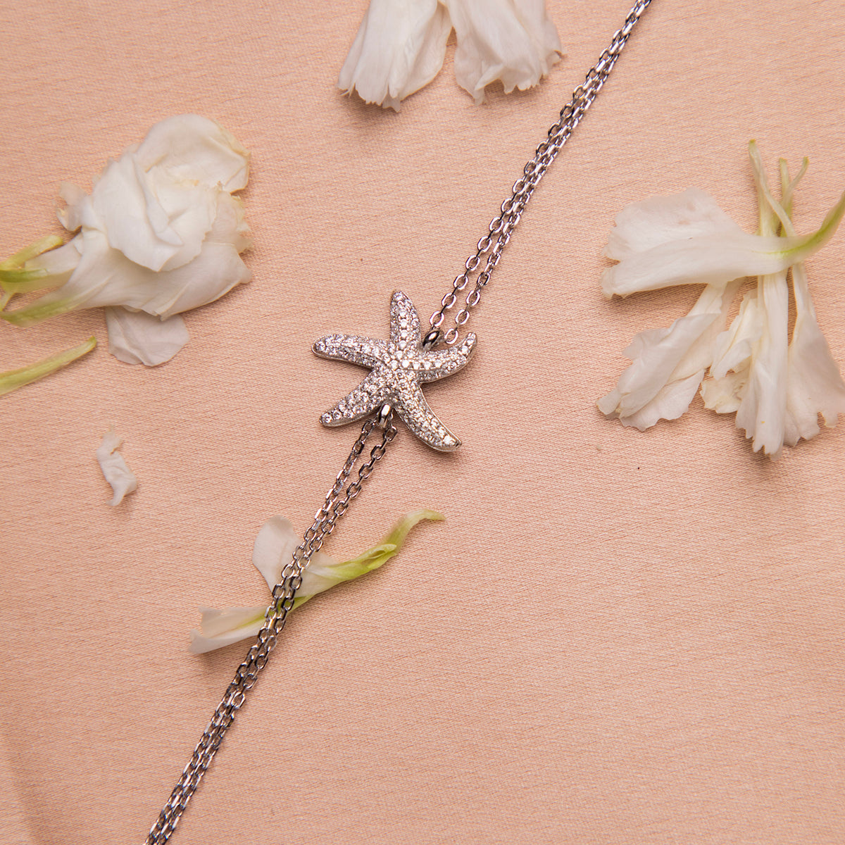 A silver bracelet featuring an embellished starfish charm, displayed on a pink background with scattered white flowers.