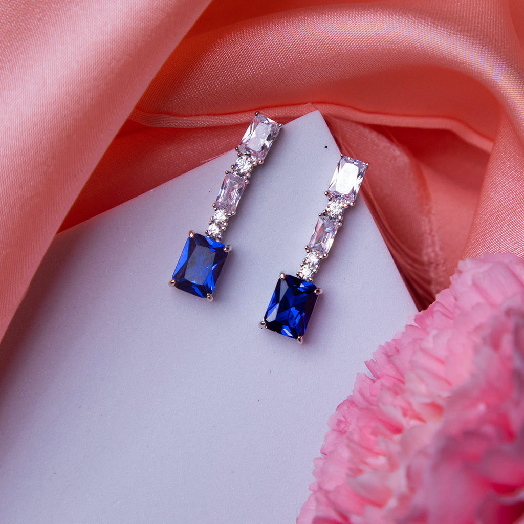A pair of emerald cut blue stone earrings with sterling silver accents, displayed on a pink satin surface.