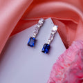A pair of emerald cut blue stone earrings with sterling silver accents, displayed on a pink satin surface.