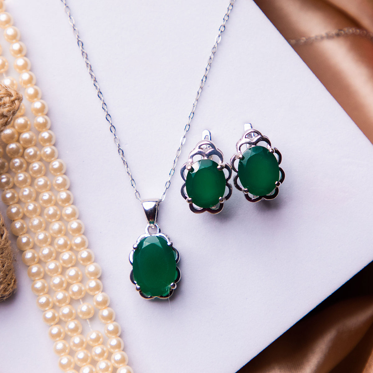 A sterling silver pendant set with emerald green stone accents, accompanied by a pair of matching earrings, displayed on a white surface with pearl accents and a velvet background.