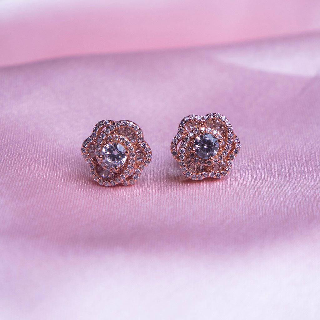 A pair of rose gold-colored evening primrose stud earrings displayed against a pink background.