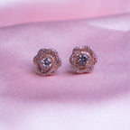 A pair of rose gold-colored evening primrose stud earrings displayed against a pink background.