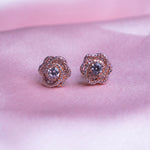 A pair of rose gold-colored evening primrose stud earrings displayed against a pink background.