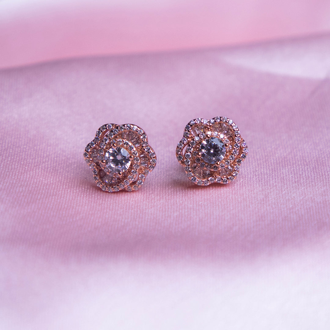 A pair of rose gold-colored evening primrose stud earrings displayed against a pink background.