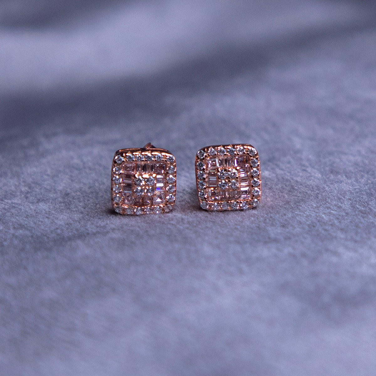 A pair of rose gold-finish stud earrings with a rhodium-based anti-tarnish coating on a textured background.