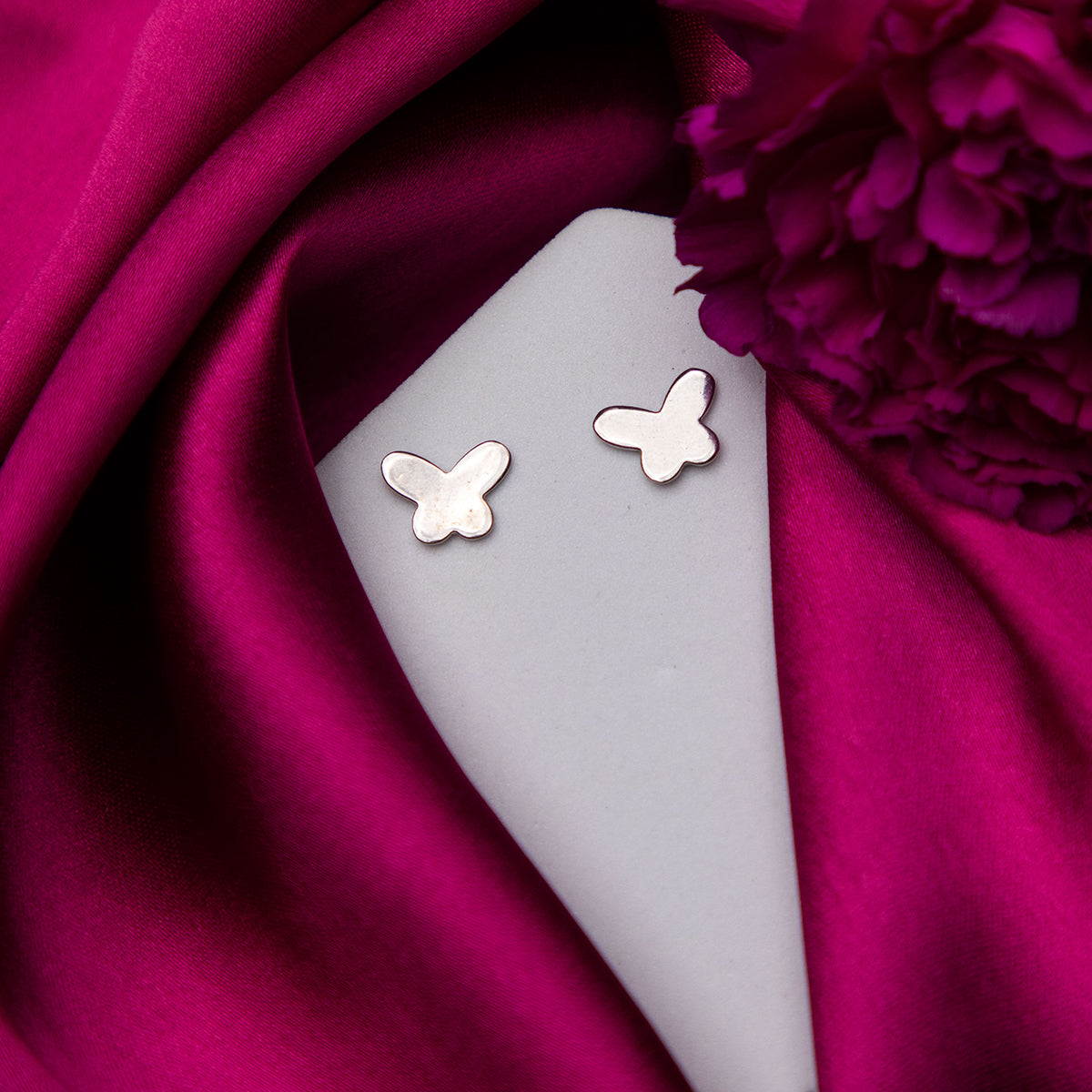 A pair of silver butterfly stud earrings on a pink fabric surface.