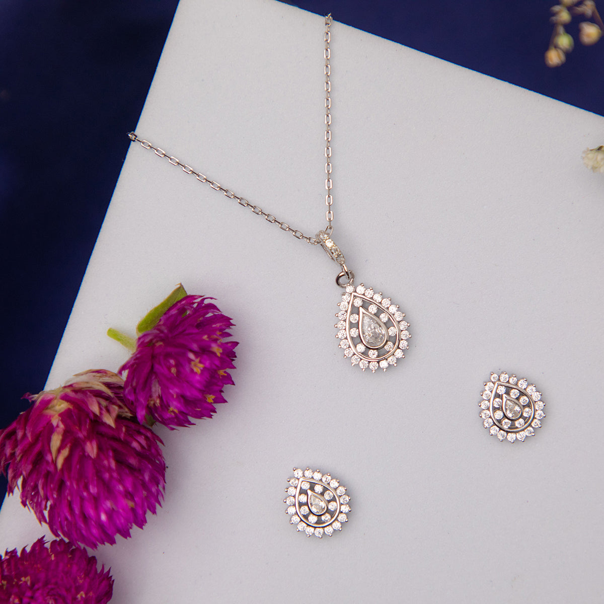 A silver pendant set with a round white stone pendant and a pair of matching earrings, displayed on a white surface with pink flowers around it.