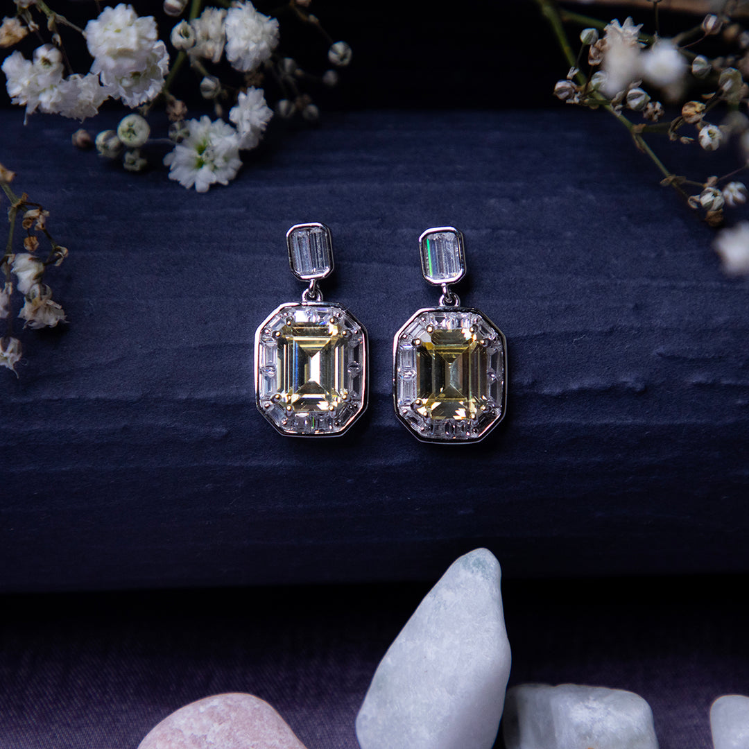 A pair of exquisite yellow gemstone earrings displayed on a textured background with floral accents and a crystal.