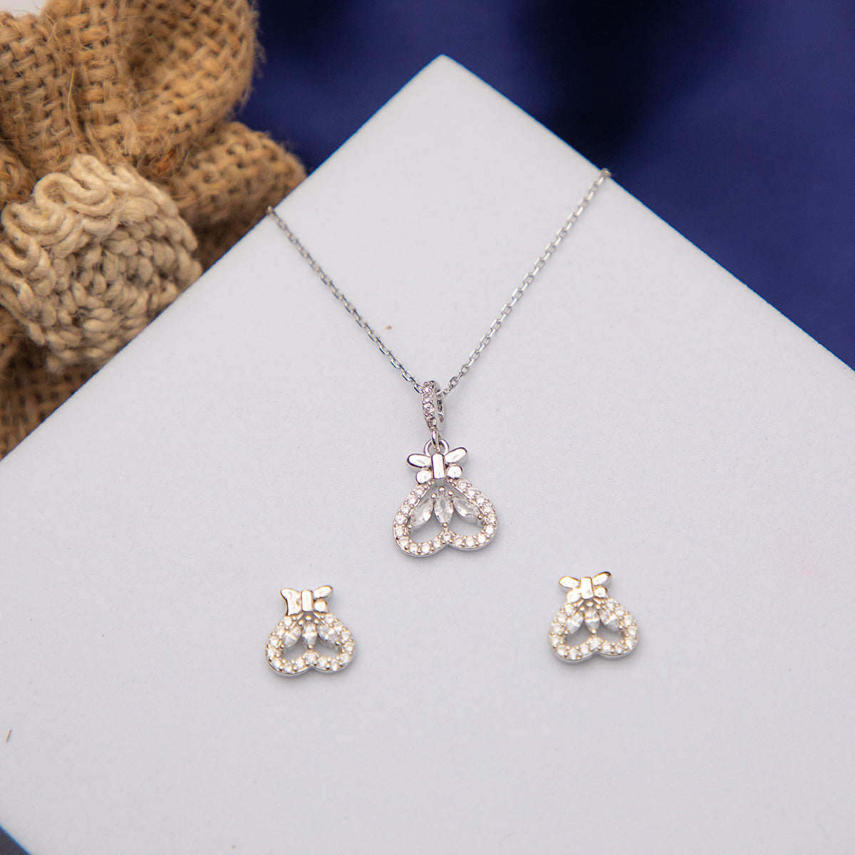 A sterling silver pendant set with a butterfly motif on an upside-down heart, accompanied by a pair of earrings, displayed on a white surface with a burlap textile in the background.
