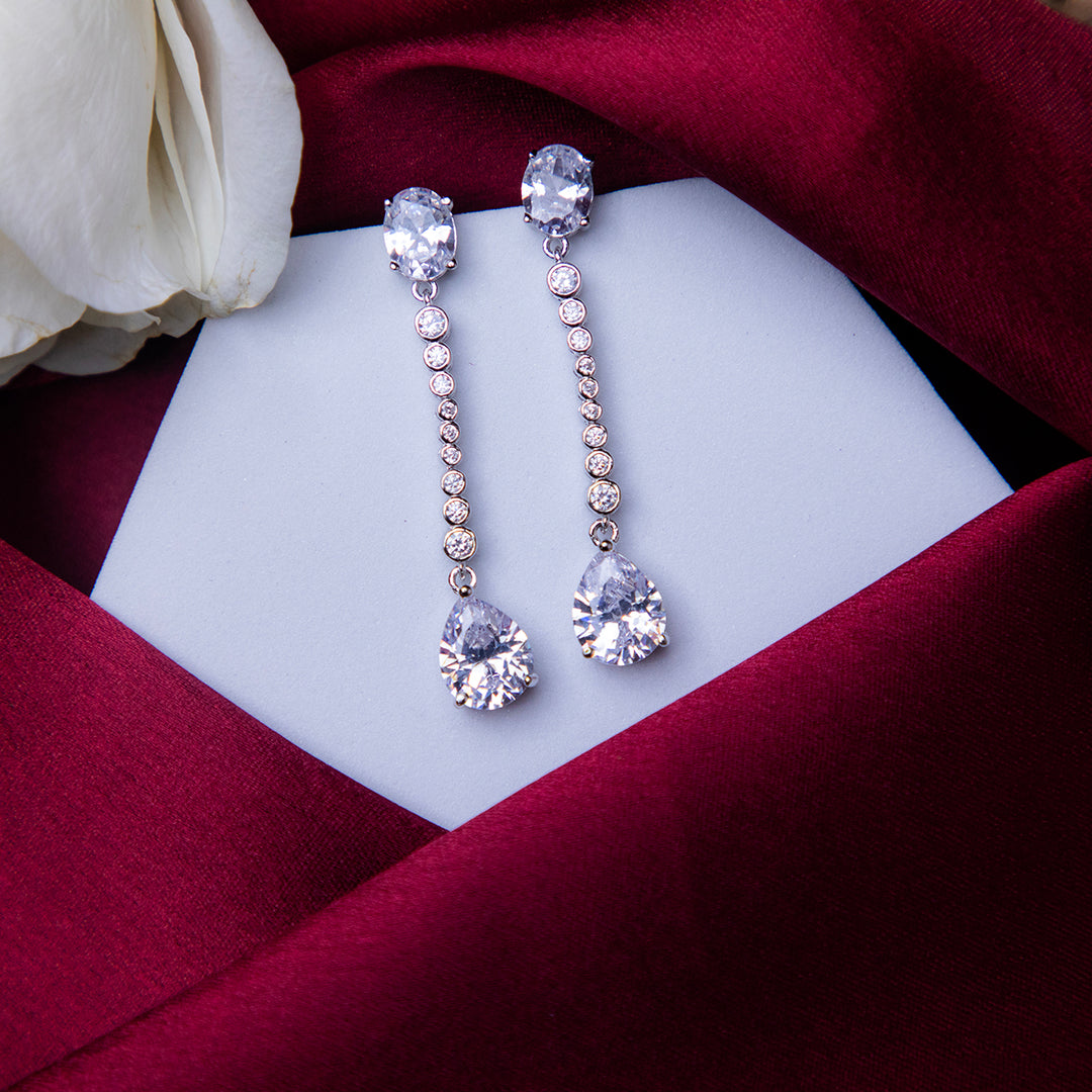 A pair of sterling silver drop earrings with white stones, displayed on a white stand with a red fabric backdrop.