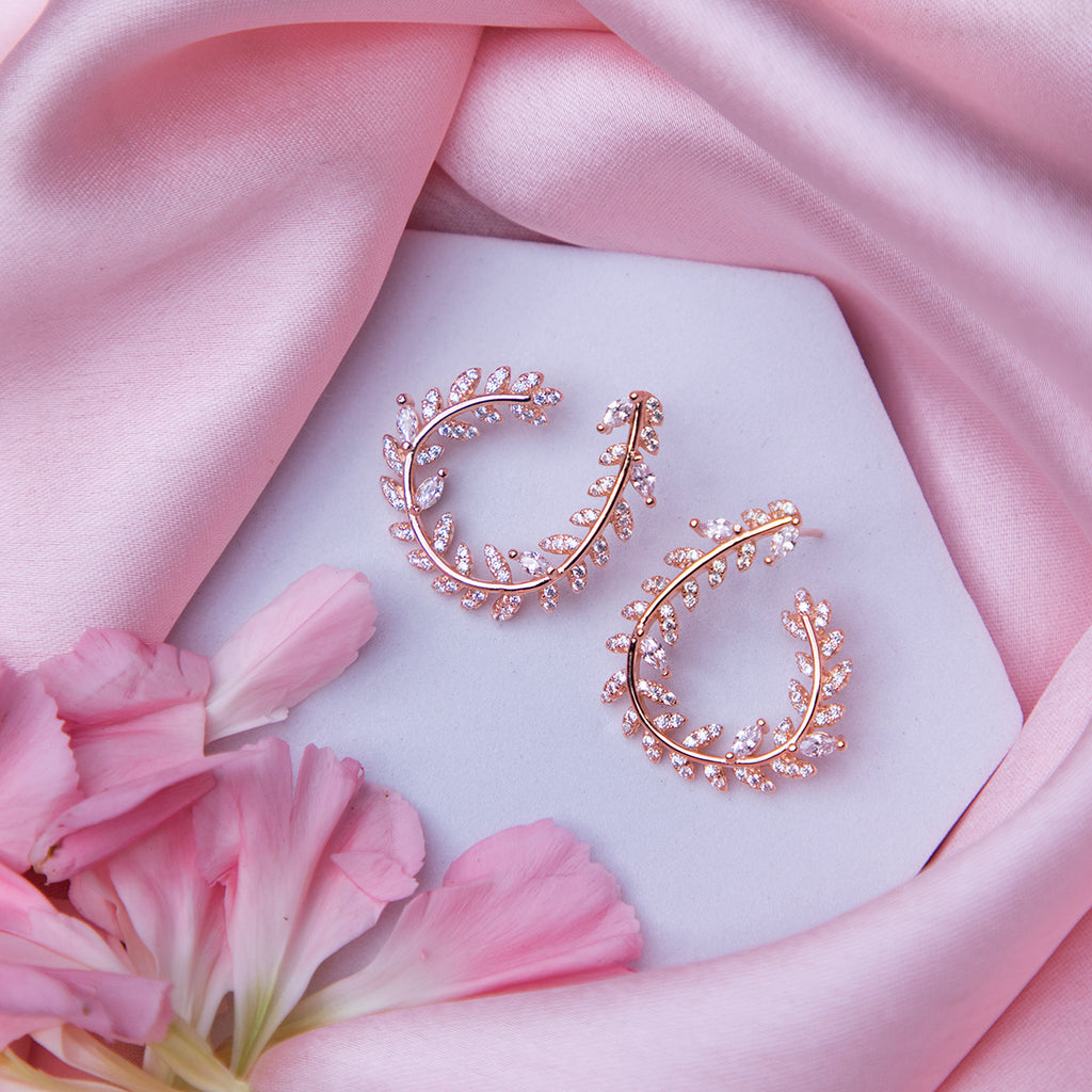 A pair of rose gold-colored fern-inspired bedazzled earrings displayed on a white surface with a pink background and a few pink flowers around.