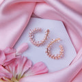 A pair of rose gold-colored fern-inspired bedazzled earrings displayed on a white surface with a pink background and a few pink flowers around.