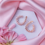 A pair of rose gold-colored fern-inspired bedazzled earrings displayed on a white surface with a pink background and a few pink flowers around.