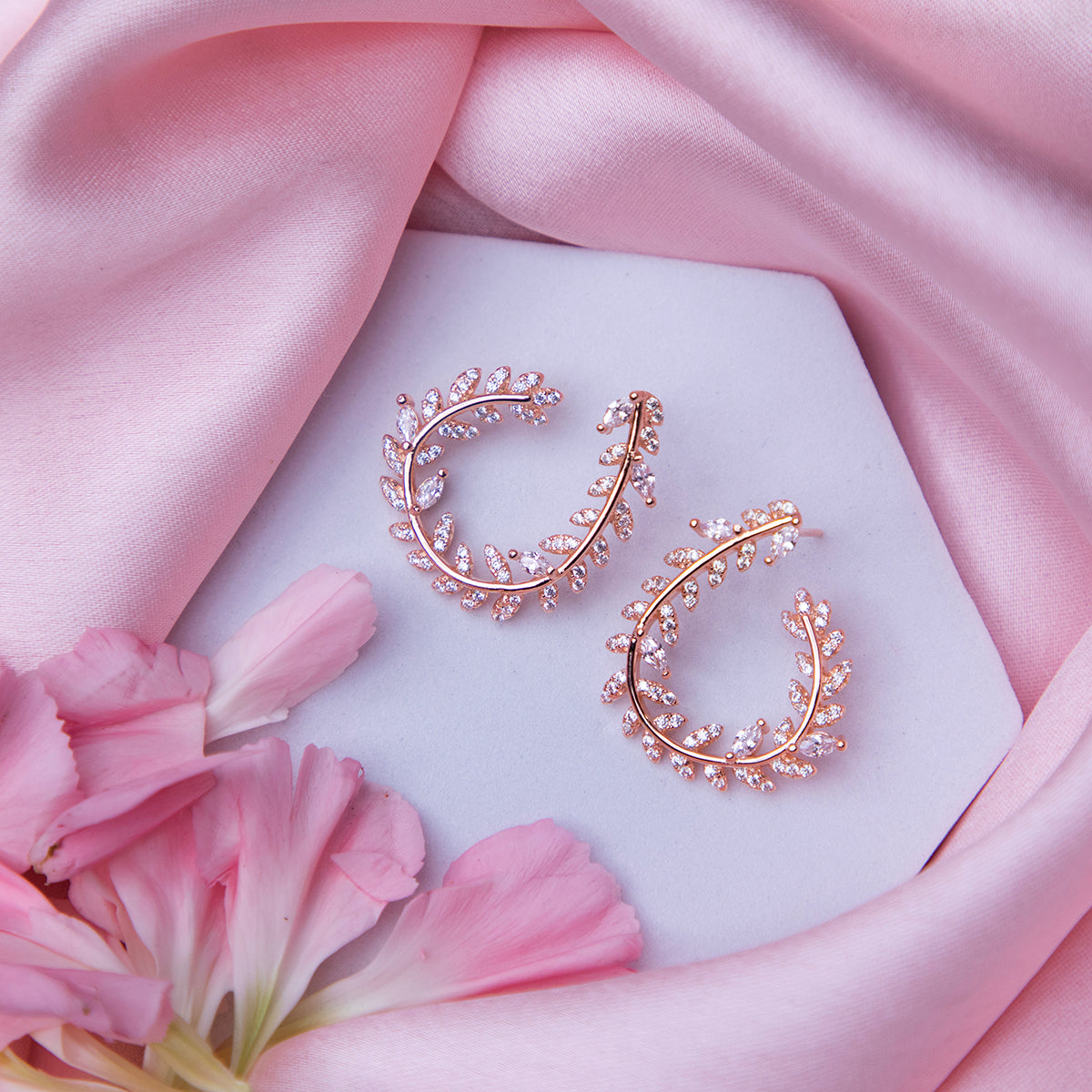 A pair of rose gold-colored fern-inspired bedazzled earrings displayed on a white surface with a pink background and a few pink flowers around.