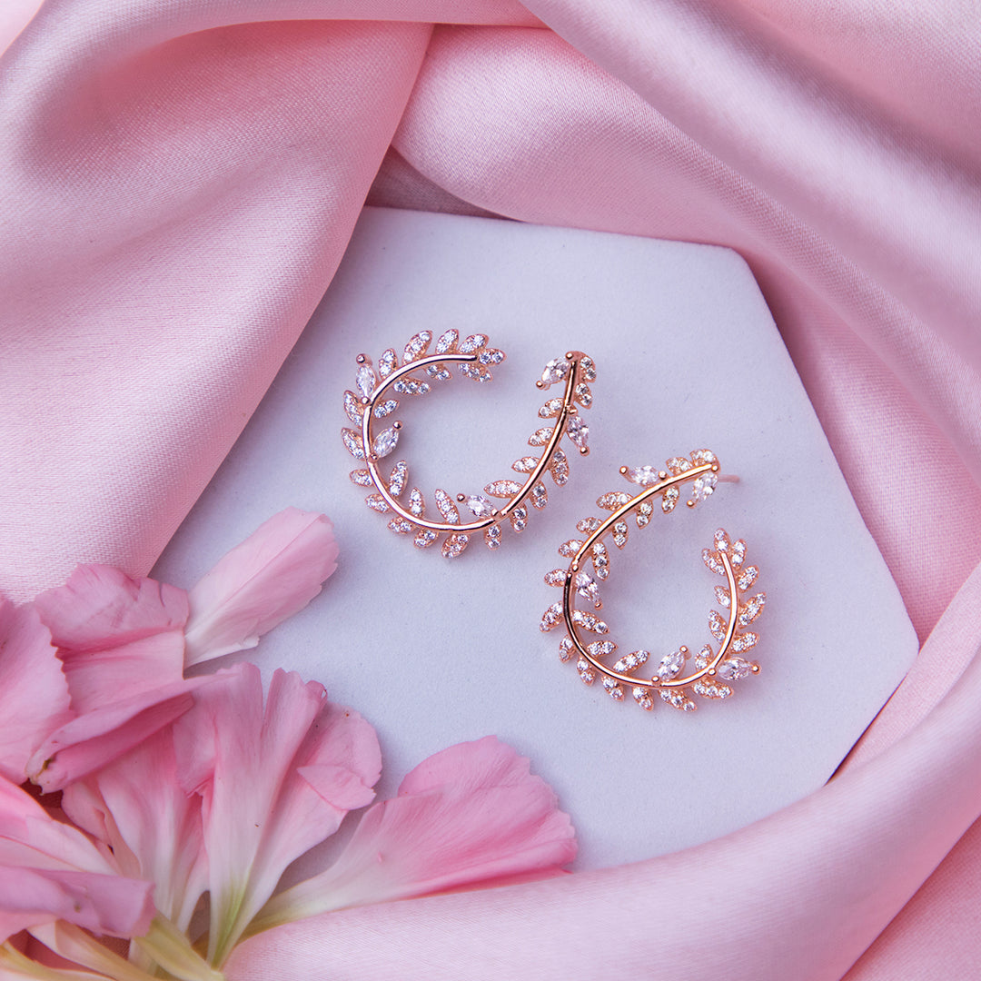 A pair of rose gold-colored fern-inspired bedazzled earrings displayed on a white surface with a pink background and a few pink flowers around.