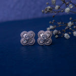 A pair of silver-colored four petal white stone stud earrings on a blue background with a decorative branch and flowers.