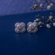 A pair of silver-colored four petal white stone stud earrings on a blue background with a decorative branch and flowers.