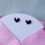 A pair of silver stud earrings with a geometric heart design, displayed on a textured pink background.