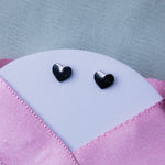 A pair of silver stud earrings with a geometric heart design, displayed on a textured pink background.
