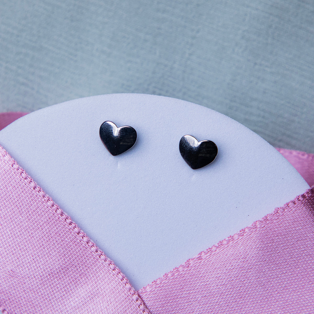 A pair of silver stud earrings with a geometric heart design, displayed on a textured pink background.