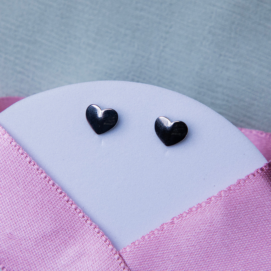 A pair of silver stud earrings with a geometric heart design, displayed on a textured pink background.