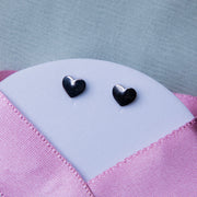 A pair of silver stud earrings with a geometric heart design, displayed on a textured pink background.