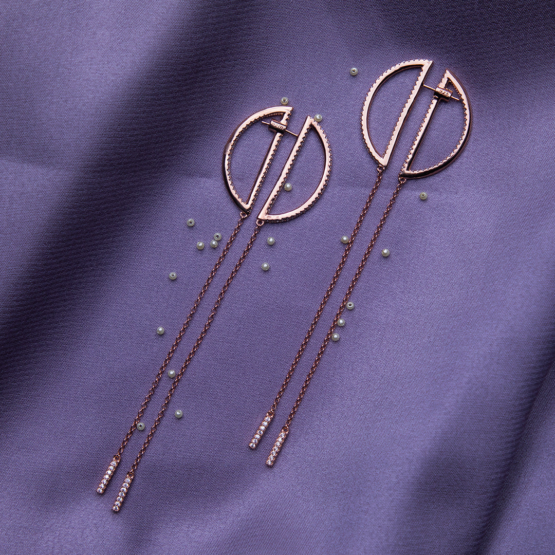 a pair of circle shaped rose gold color silver earrings dangler laid over a purple fabric with pearls sprinkled around it