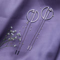 A pair of geometric silver chain earrings laid out on a purple fabric surface, accompanied by small decorative flowers.