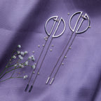 A pair of geometric silver chain earrings laid out on a purple fabric surface, accompanied by small decorative flowers.