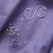A pair of geometric silver chain earrings laid out on a purple fabric surface, accompanied by small decorative flowers.