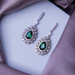 A pair of glittering green gemstone statement earrings laid on a silky purple fabric.