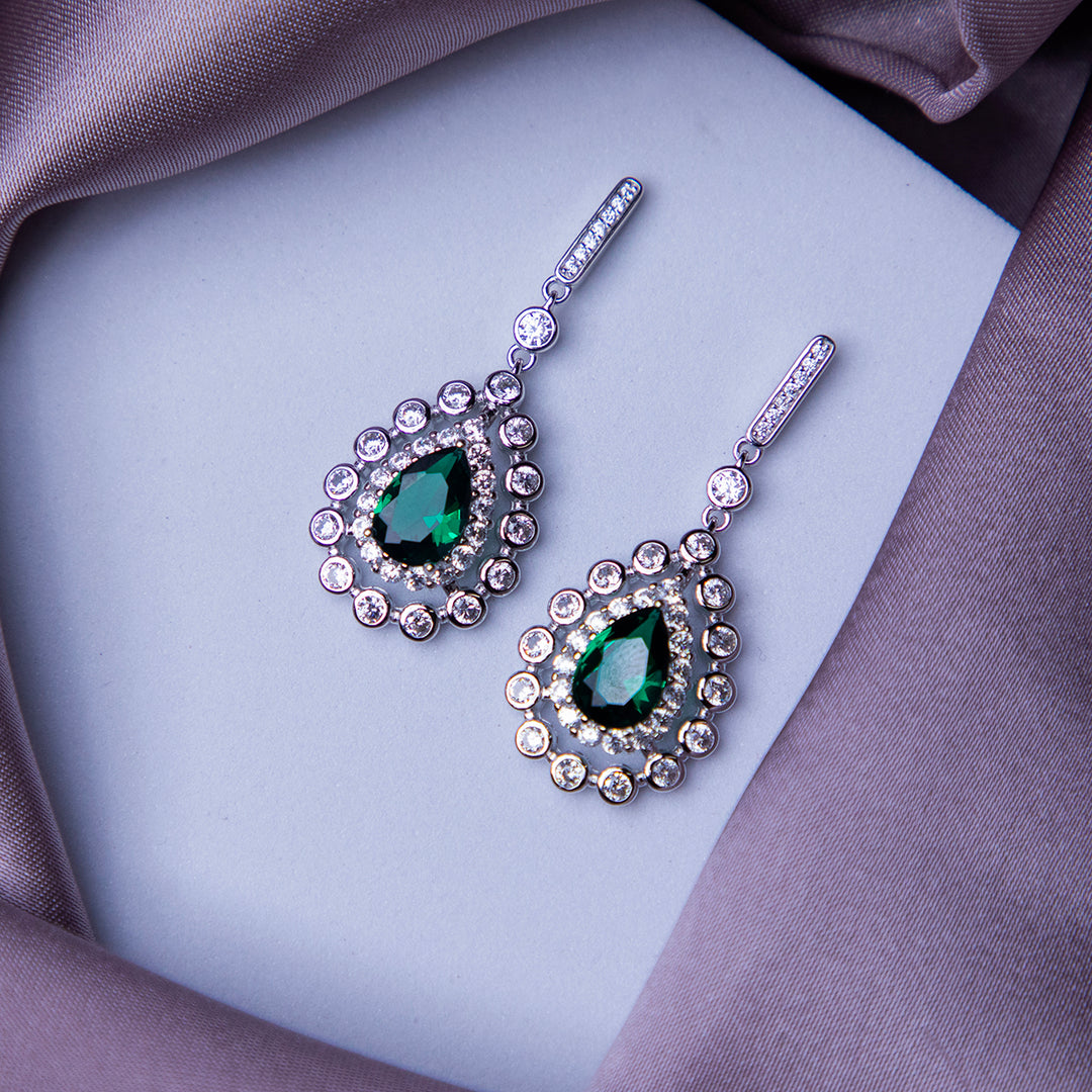 A pair of glittering green gemstone statement earrings laid on a silky purple fabric.