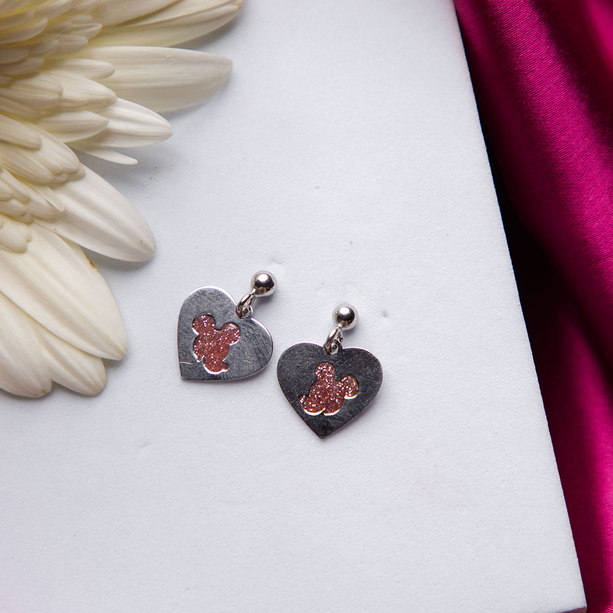 A pair of silver-colored Mickey Mouse stud earrings placed on a white surface with a pink and white flower background.
