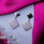 A pair of silver geometric drop earrings with a rhodium-based anti-tarnish coating, displayed on a surface with a pink scarf and white flowers in the background.