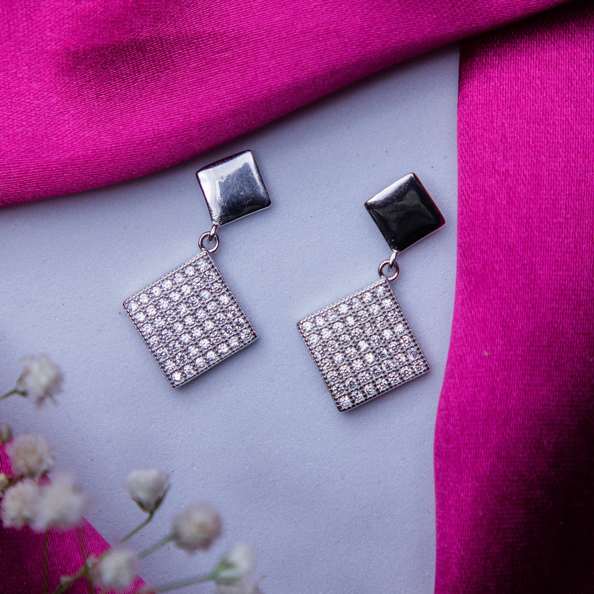 A pair of silver geometric drop earrings with a rhodium-based anti-tarnish coating, displayed on a surface with a pink scarf and white flowers in the background.