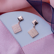 A pair of rose gold-color drop earrings with a geometric pattern, placed on a surface with a pink fabric backdrop.