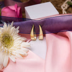 A pair of gold-colored silver hoop earrings with a geometric design, displayed on a pink fabric surface next to a white flower.