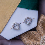 A pair of silver stud earrings with a geometric pattern, displayed on a white card, with a yellow textured background and a burlap fabric flower next to it.